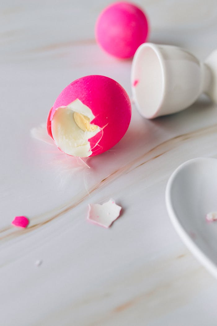 Close-up of a vibrant pink colored broken eggshell next to an egg cup on a white marble surface.